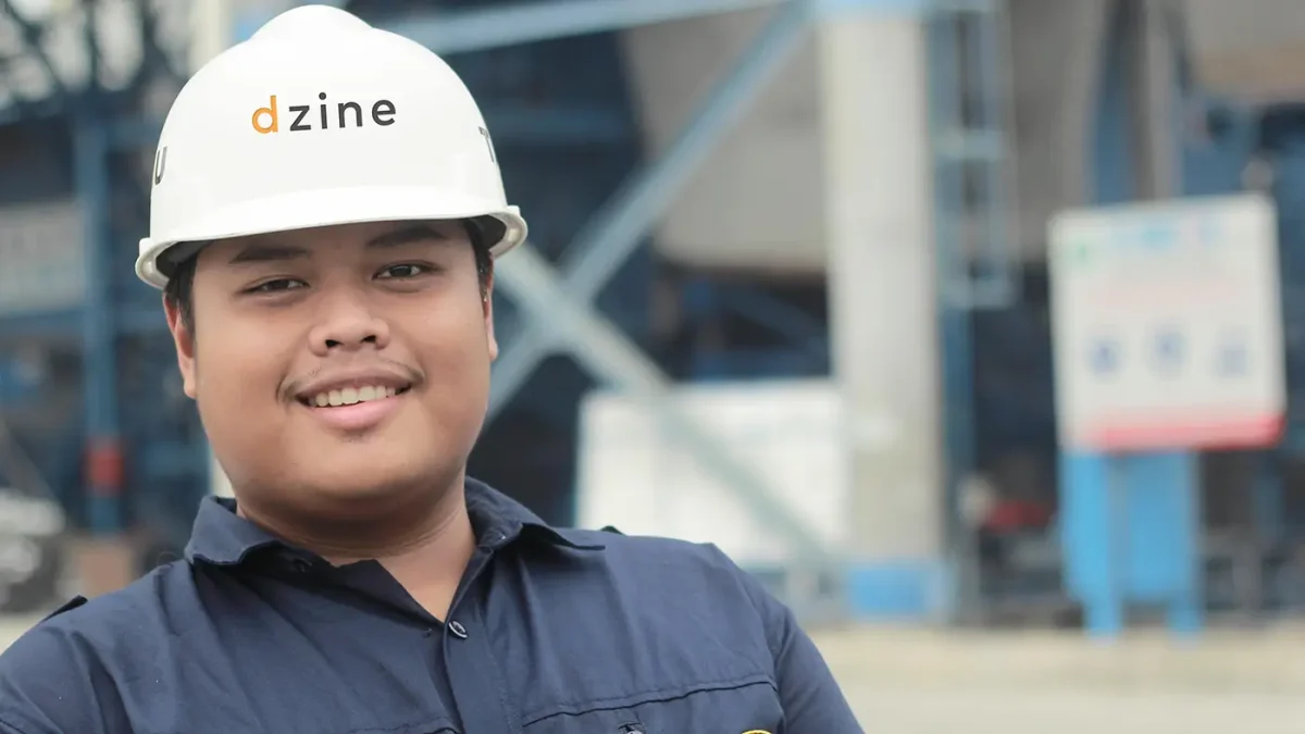 Person in a hard hat and uniform smiling outside near industrial equipment.